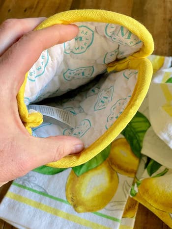 Hand holding an insulated oven mitt with citrus design, adjacent to matching lemon-patterned room towels connected a woody surface