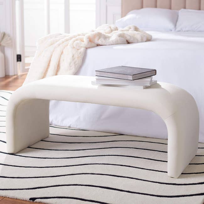 Curved modern bench with books on it, positioned at the foot of a bed