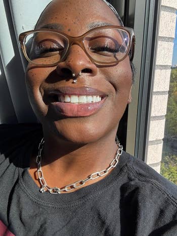 Person wearing glasses and a chain necklace smiles while standing by a window