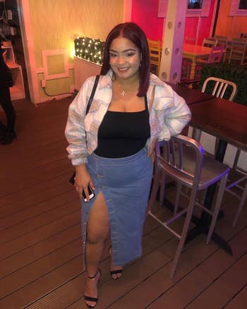 A pistillate   stands successful  a casual setting, wearing a checkered jacket, achromatic  top, and denim skirt with a precocious   slit. She smiles, holding a phone