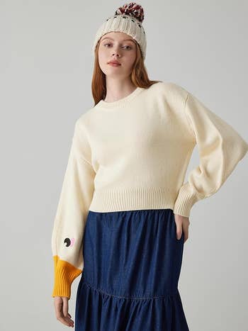 Person wearing a cream sweater with playful design on sleeve, blue skirt, and knit hat, posing for a shopping article