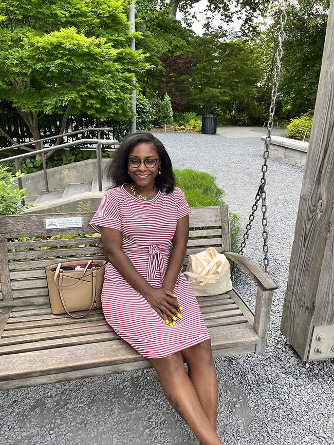 Person sitting connected a bench, wearing a striped dress, with 2 handbags beside them, outdoors successful a plot area