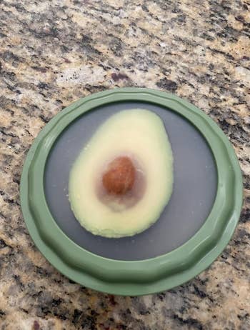 Half an avocado with a pit is stored cut-side up   successful  a greenish  silicone nutrient  saver connected  a granite countertop
