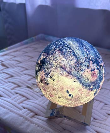 Glowing planet-like lamp on a small wooden stand, set on a woven surface, showcasing an artistic and celestial design. Ideal for home decor