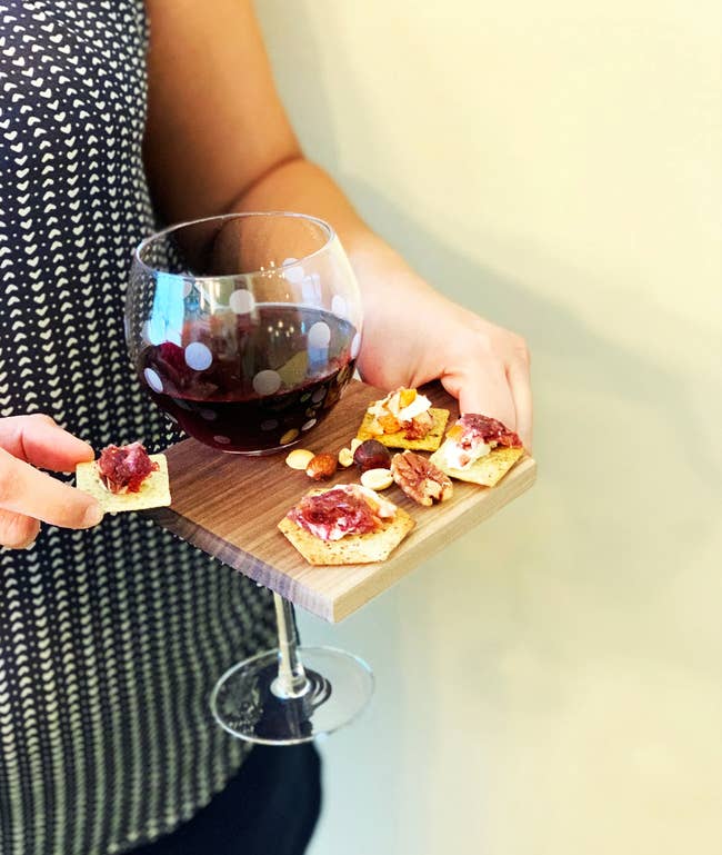A person holding the tray with one hand filled with crackers and a glass of wine atop while they're holding a cracker in the other hand