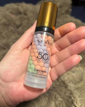 Hand holding a vessel of SADOOER SPF 50 PA+++ sunscreen with three-color concealer pick beads