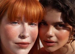 Models wearing orange blush on cheeks and lips, one with fair skin and freckles and the other with medium olive skin