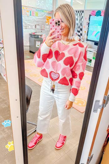 reviewer in a heart-patterned sweater and white pants taking a mirror selfie in a room with visible floor stickers and eyewear displays