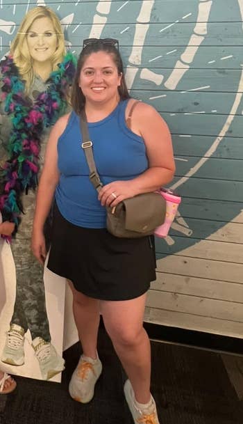 Person successful casual attire with a crossbody container poses indoors adjacent to a cardboard cutout wearing a colorful feather boa