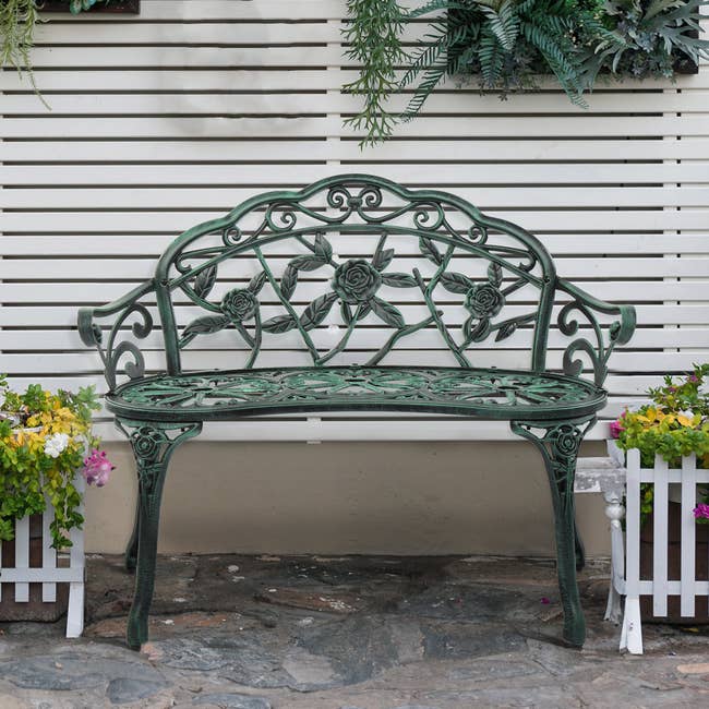 Decorative metallic plot seat with floral design, flanked by potted plants against a achromatic slatted wall