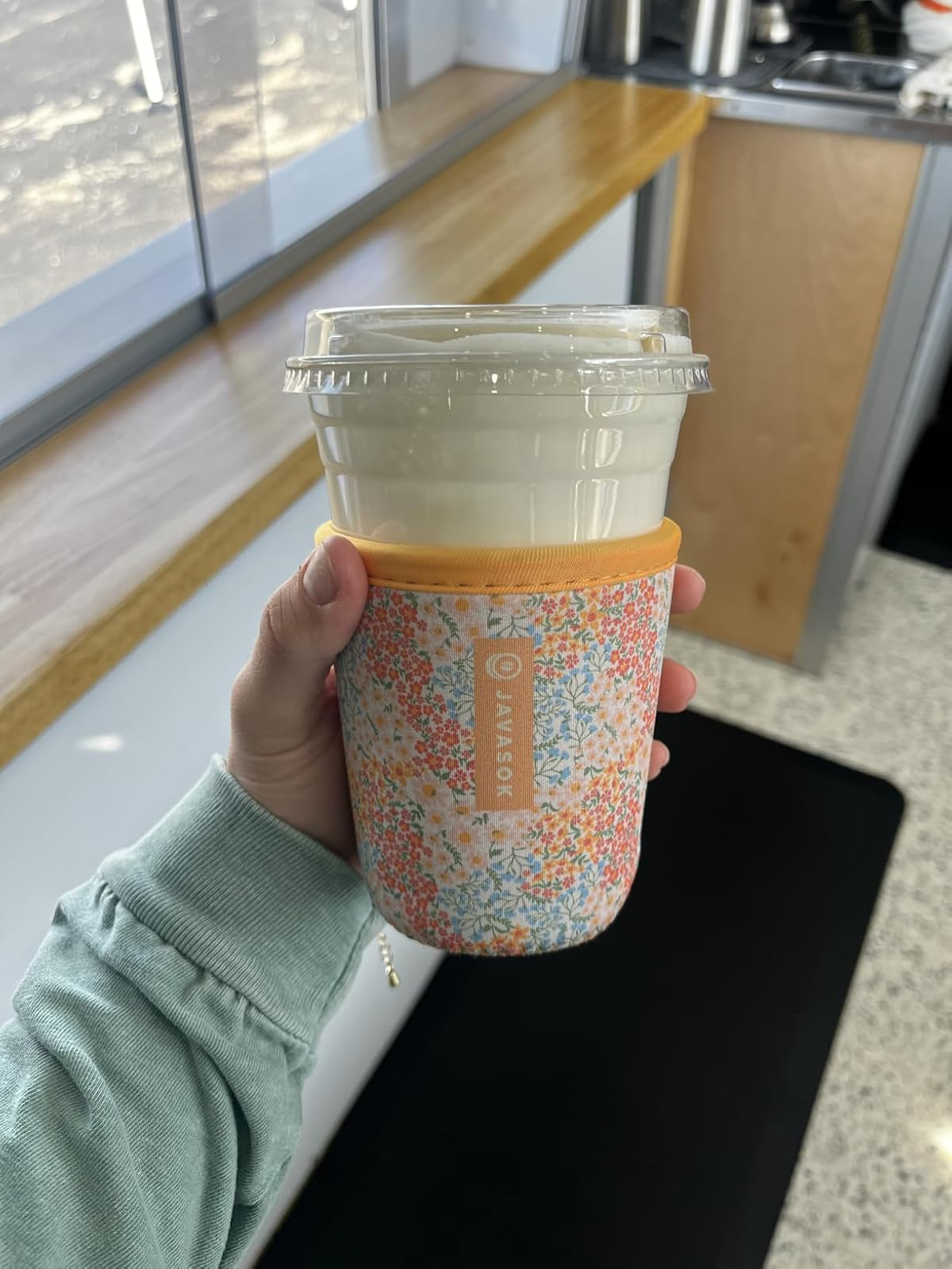 Hand holding a drink with a clear lid in a floral insulated sleeve, inside a cozy café setting