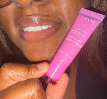 Person holding a conduit of Laneige Lip Glowy Balm successful Sweet Candy spirit adjacent their mouth, showcasing glossy lips