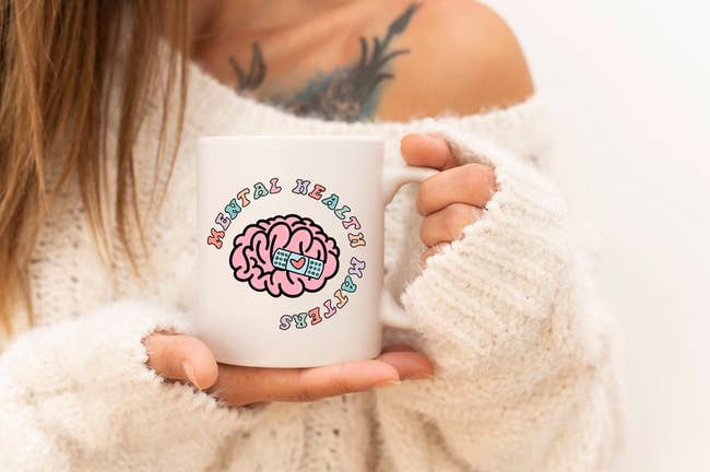 model holding white mug with pastel writing and an illustration of a brain with a heart bandage on it
