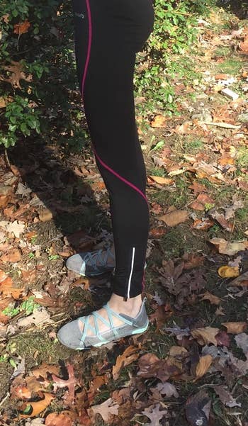 Reviewer wears pair of black thermal leggings with a pink stripe going down the legs and zippers near the ankles with running shoes while stepping on a pile of leaves