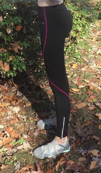 Reviewer wears pair of black thermal leggings with a pink stripe going down the legs and zippers near the ankles with running shoes while stepping on a pile of leaves