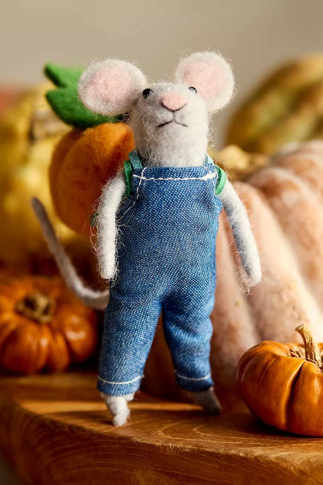 A felt rodent figurine wearing denim overalls stands surrounded by tiny pumpkins, suggesting a cozy, autumn-themed decoration