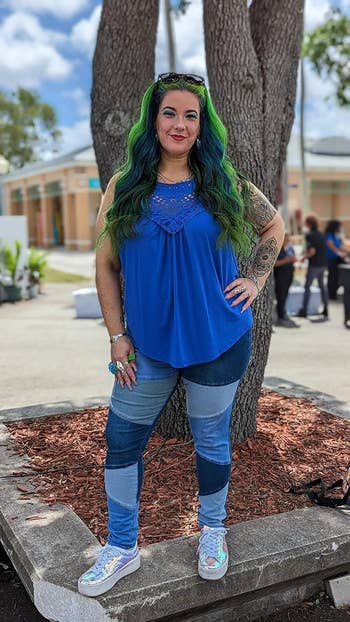 A reviewer with long, green-tinted hair, wearing a blue sleeveless top and patchwork jeans, stands outside.