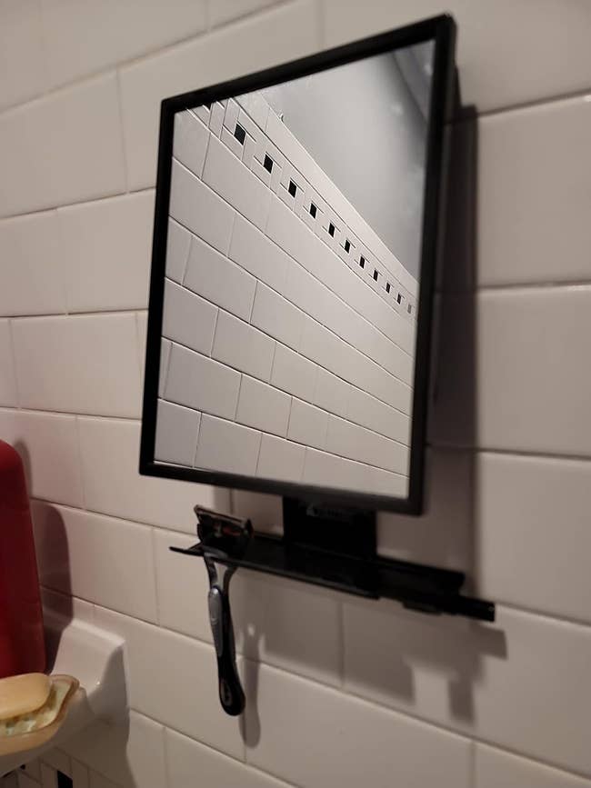 Mirror mounted on tiled wall reflecting tiles, with a razor on a ledge below it