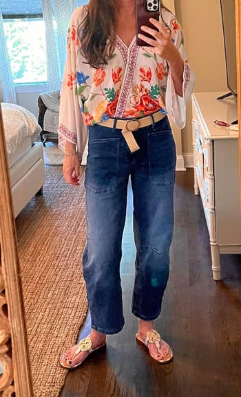 Seseorang mengambil selfie cermin mengenakan blus bunga, celana jeans biru, dan sandal dengan desain cut-out dekoratif