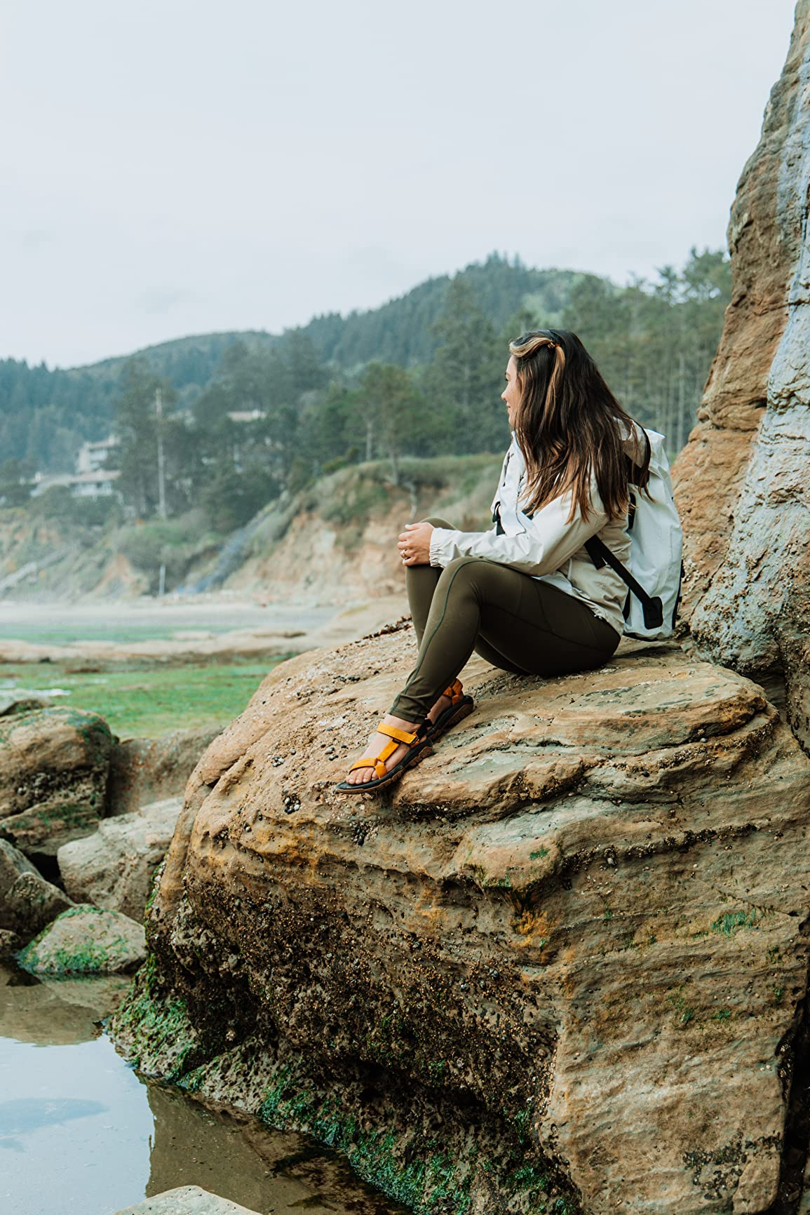 Person sitting connected  a rocky beach, wearing a casual outdoor outfit with a backpack, looking towards the ocean