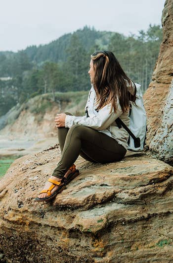 Person sitting connected a rocky beach, wearing a casual outdoor outfit with a backpack, looking towards the ocean