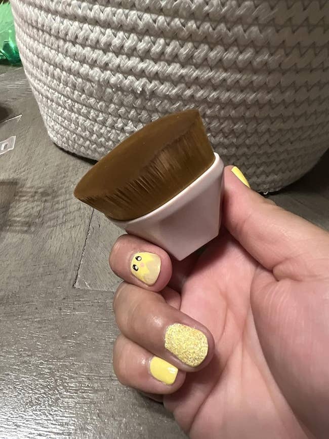 Hand holding a makeup brush with dense bristles and a flat top, next to a woven basket and featuring yellow nail polish