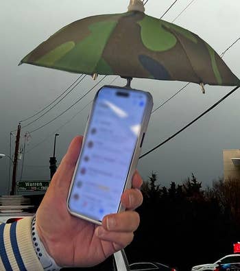 Person holding a smartphone under a small camouflage-patterned umbrella to shield it from a stormy day