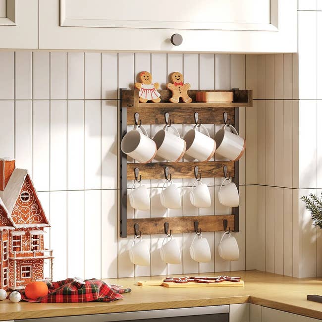 Wooden mug rack on a kitchen wall holding eight white mugs, with gingerbread figures above. Nearby, a gingerbread house and holiday decor are displayed
