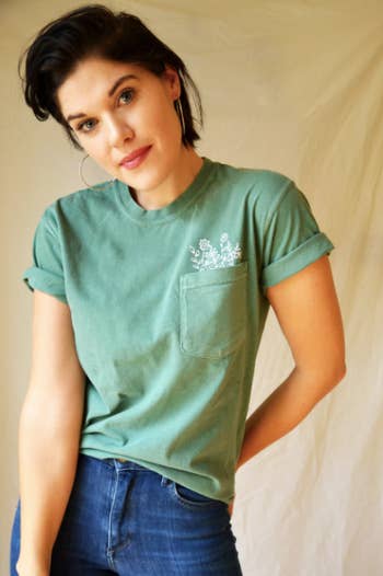 model in green short sleeve tee with white illustration of flowers peeking out of pocket