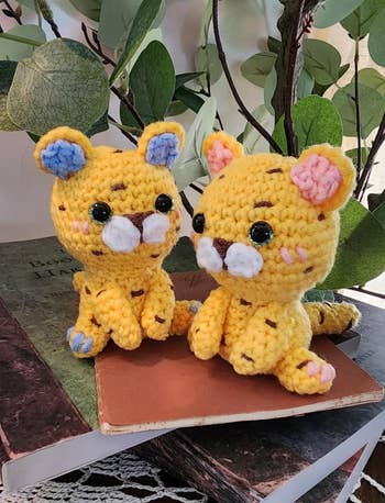 Two yellow crocheted cheetah dolls with large eyes, sitting on books with leaves in the background