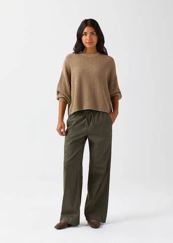 Person wearing a loose, ribbed sweater and wide-leg trousers, lasting against a plain backdrop