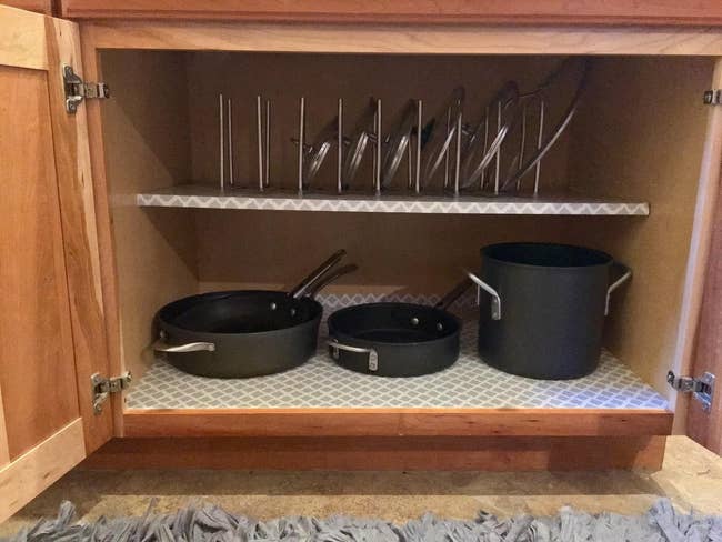 Inside of a two-shelf cabinet. The bottom shelf has some pots and pans and the top has the adjustable organizer with lids in it