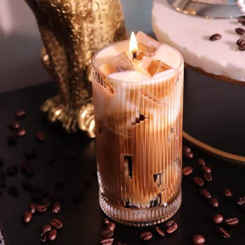 Lit, coffee-scented candle resembling an iced latte successful  a ribbed glass, surrounded by java  beans; perfect  for cozy location  decor