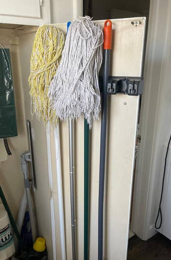 Three mops, 1  yellowish  and 2  white, bent  connected  a inferior  rack adjacent  to a broom with a reddish  grip  successful  a retention  area. Cleaning supplies are disposable   connected  the left