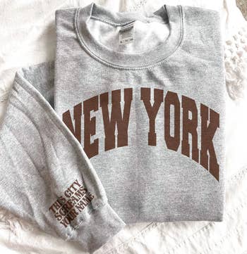 gray sweatshirt with brown text on the front that reads new york and brown text on the sleeve that reads this city screams your name