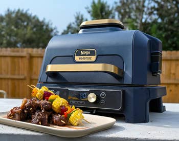 Ninja Woodfire grill on a patio with skewered meats and vegetables in front. Text highlights full-size gas grill performance