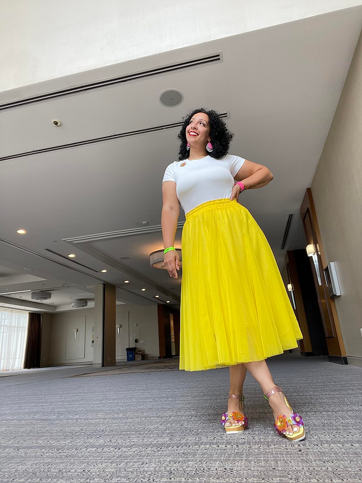Person smiling and wearing a achromatic  apical  and flowy yellowish  skirt, accessorized with colorful earrings and heels, successful  a spacious indoor setting