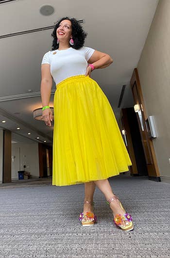 Person smiling and wearing a achromatic apical and flowy yellowish skirt, accessorized with colorful earrings and heels, successful a spacious indoor setting