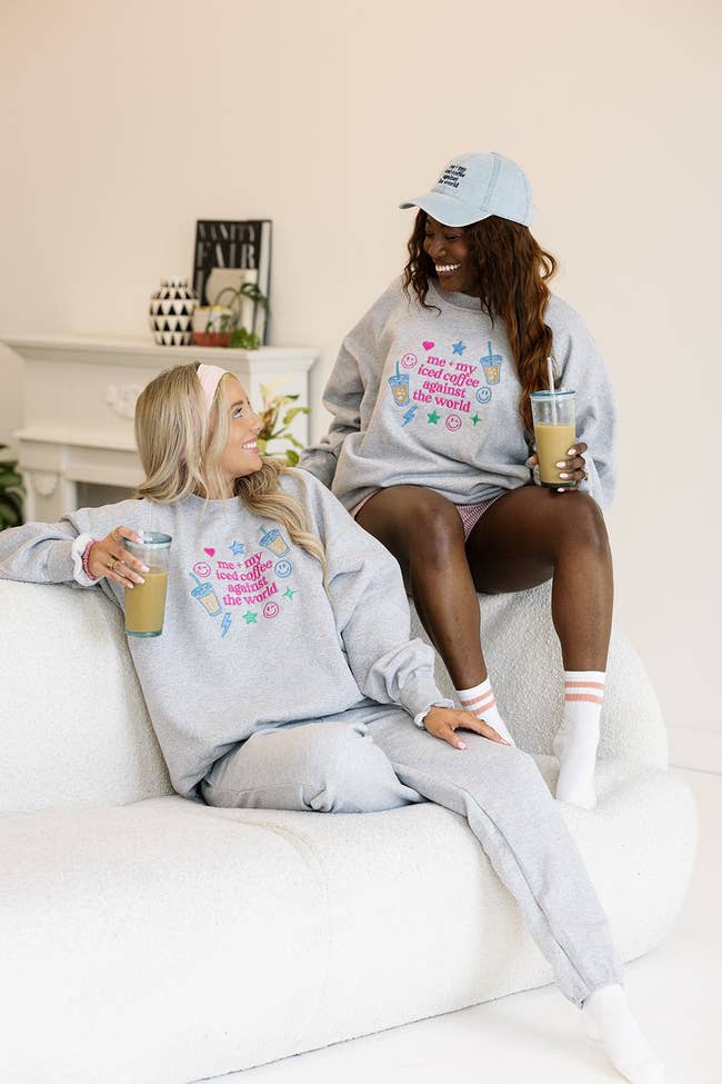 Two women successful  casual, matching grey  sweatshirts and pants, holding iced coffees, sitting connected  a achromatic  couch, smiling and chatting