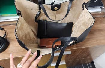 Open handbag with a tiny achromatic pouch and tan wallet inside. Person's manicured manus reaching for items