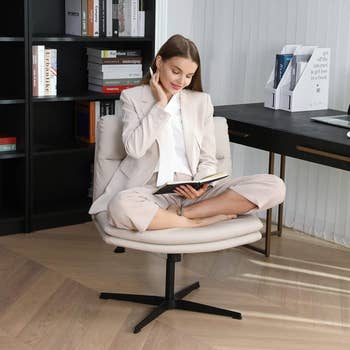 Woman successful  tailored suit   sits cross-legged connected  chair, speechmaking  a publication  successful  a stylish, modern   bureau   mounting  with bookshelves and a table  successful  the background