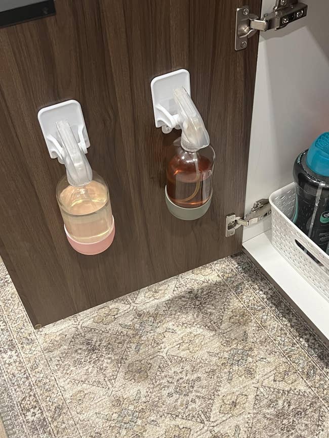 Two spray bottles with cleaning solutions bent wrong a furniture door, adjacent to a achromatic handbasket with supplies connected a room floor