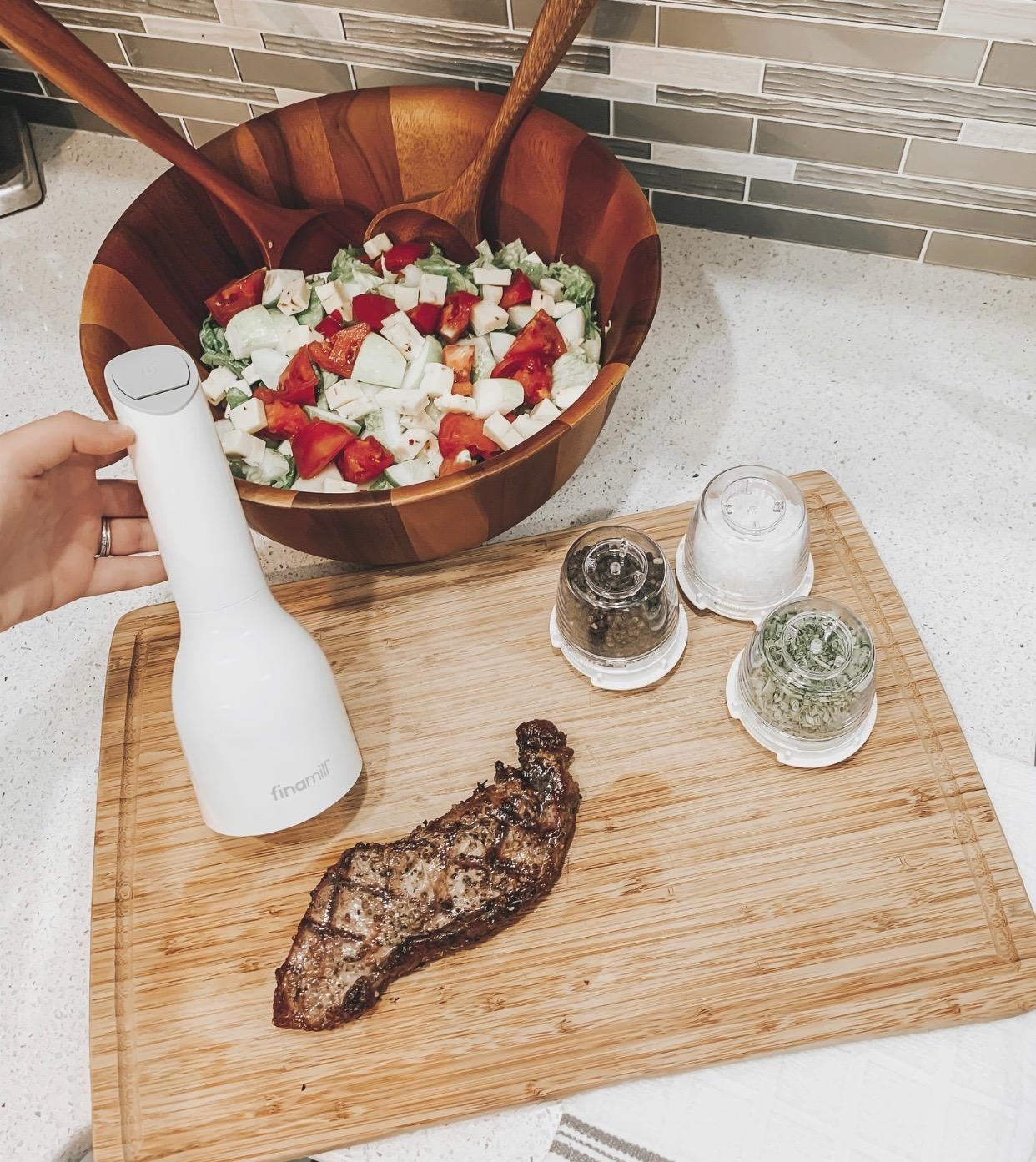 image of reviewer holding the white automatic spice grinder over a cutting board with a steak on it