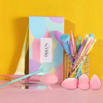 Assorted makeup brushes in a cup with beauty sponges and pastel-colored packaging on a yellow background