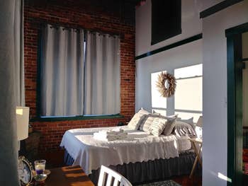 A well-lit bedroom with a neatly made bed, sheer curtains, and exposed brick walls, showcasing cozy decor