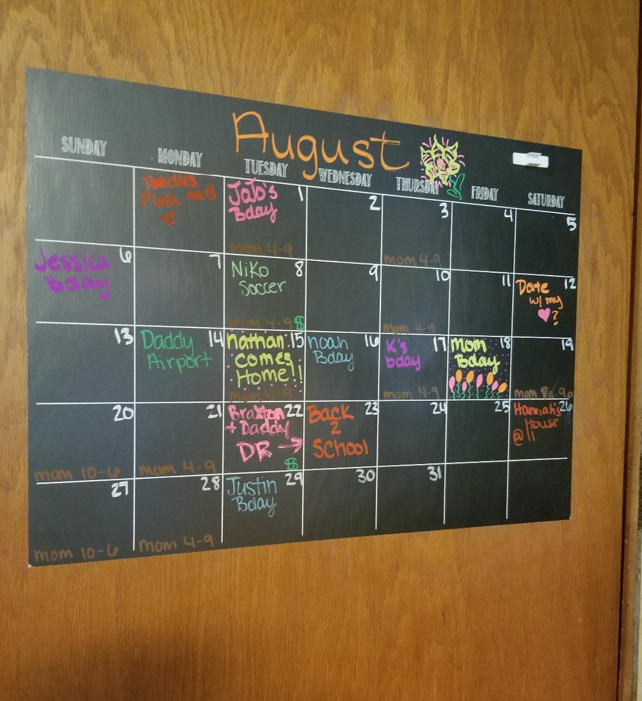 A black calendar with colorful pen writing dates and events on it adhered to a wall 