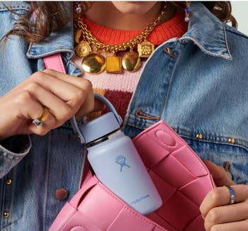Person holding a tiny  Hydro Flask and pinkish  woven bag, wearing a denim overgarment   and layered necklaces