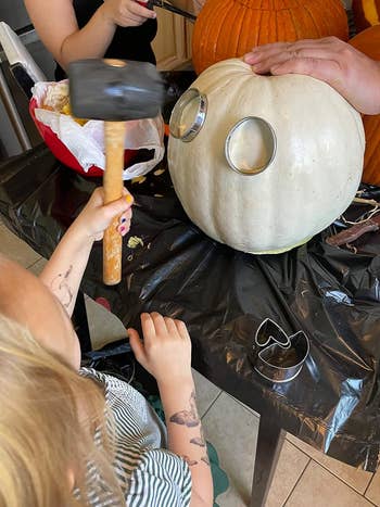 A child using a rubber mallet to hammer a stainless steel cutout into a white pumpkin