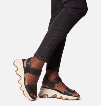 Person wearing black strapped sandals with thick, wavy soles paired with dark jeans, showcasing a trendy footwear choice for casual outfits