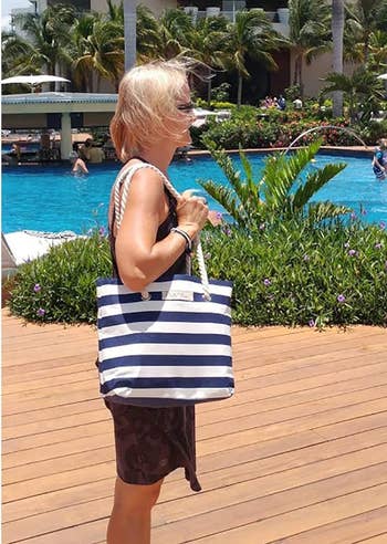A person carrying a striped tote bag stands near a pool with people swimming and lounging. A multi-story building with palm trees is in the background